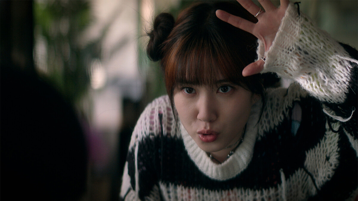 Young woman indoors with plants in the background, wearing a black and white sweater with frayed sleeves, hair in two buns, raising her hand and making a playful expression, as if acting or interacting animatedly.