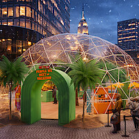 People line up outside a large transparent dome with palm trees and vacation-themed decor in a city plaza at night, surrounded by tall buildings and light snowfall.