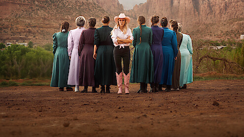 A group of women in long, modest dresses stand in a dirt area, backs to the camera, with one woman in a cowboy hat, white shirt, black pants, and pink boots facing forward against a backdrop of mountains and greenery.