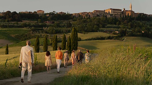 A group of people walk down a country path through green fields at sunset, heading toward a distant hilltop village, evoking a peaceful and warm atmosphere.