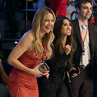 Three people dressed formally stand indoors at an event, two women in front appear excited and cheering, holding phones, while a man in a suit stands beside them, all wearing lanyards, with a blurred audience in the background.