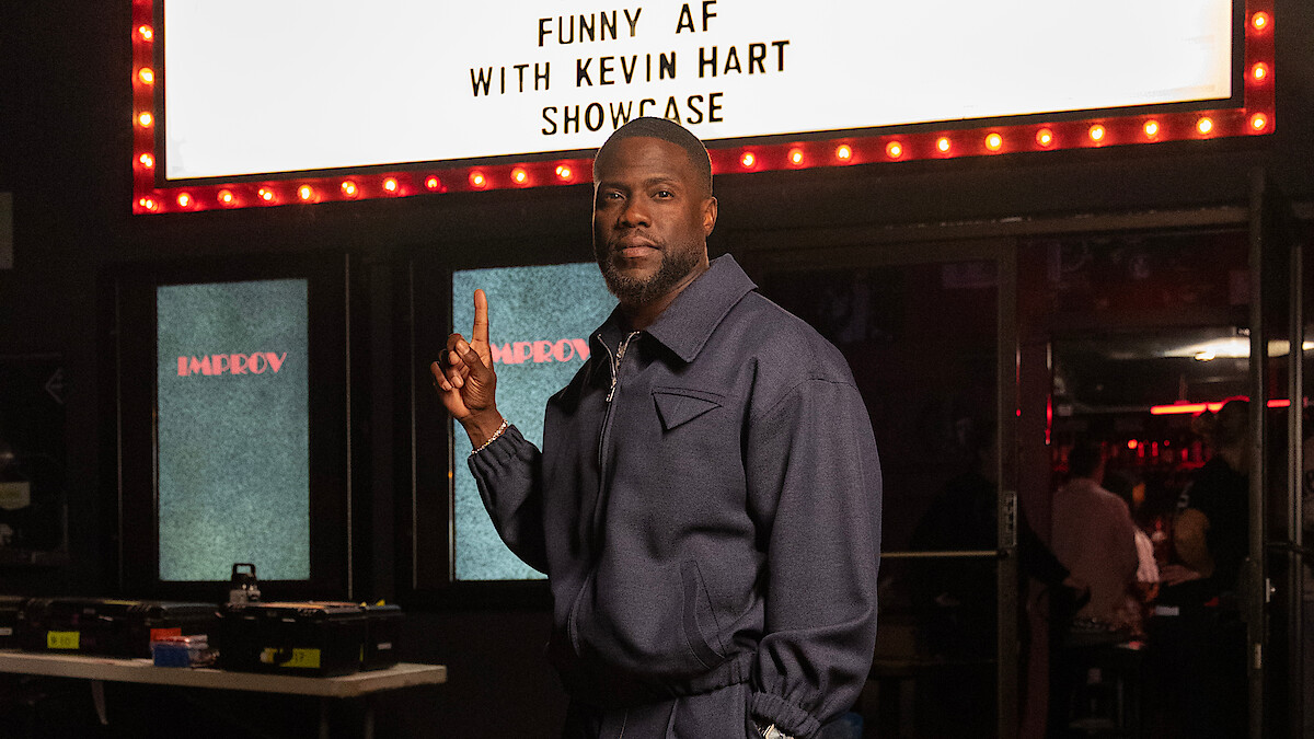 Kevin Hart in a blue jacket stands inside a comedy club, pointing upward at a lit marquee sign that reads “Funny AF with Kevin Hart Showcase,” with tables, equipment, and an audience in the background.