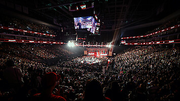 Large, packed arena hosting a WWE wrestling event with bright lights, digital screens, and enthusiastic crowd surrounding a central ring under the arena’s high ceiling.