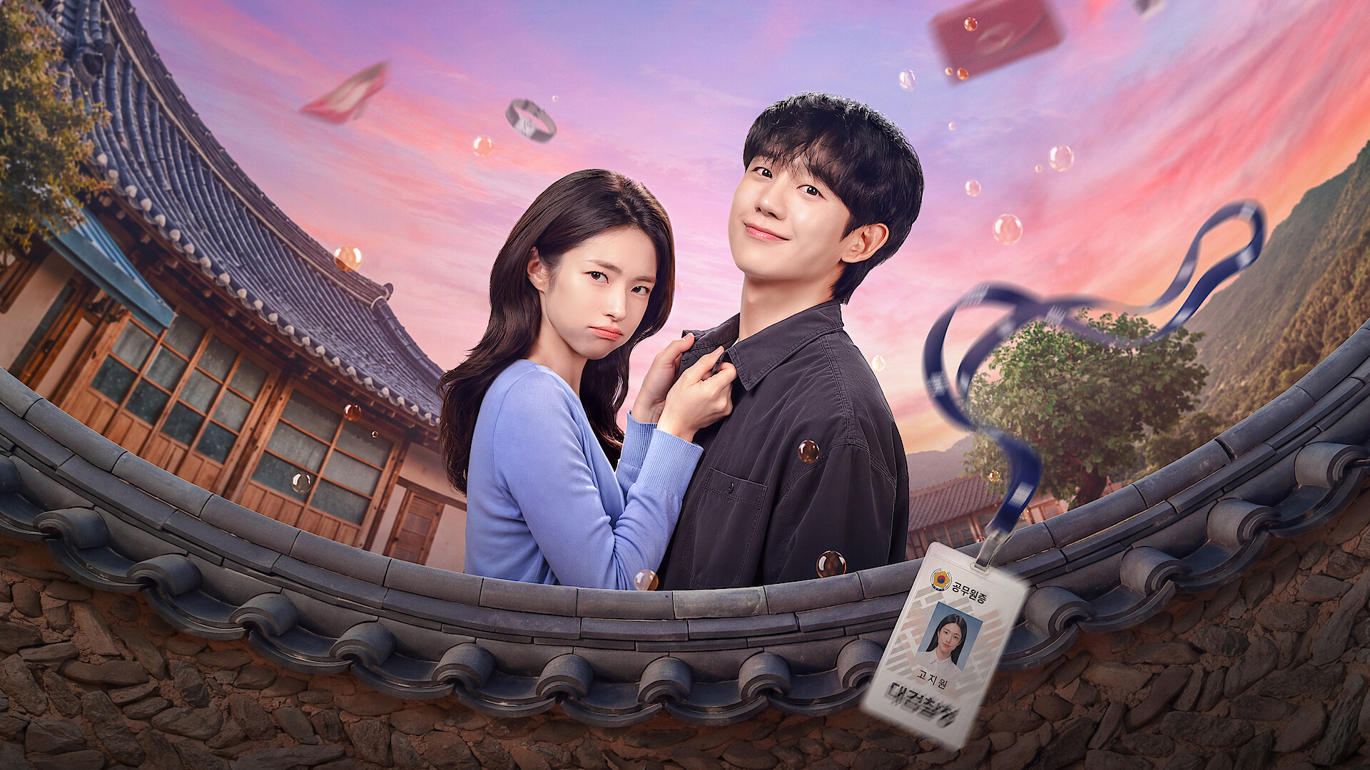 Two young adults stand close together, one holding the other's collar, in a traditional Korean courtyard with mountains at sunset and floating identification cards and books in the air, suggesting a dramatic or magical setting.
