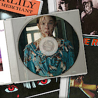 A CD case with an image of a young woman in a floral dress, covered in blood, in a dimly lit room. The CD is on top of various music album covers featuring bold colors and graphic artwork.