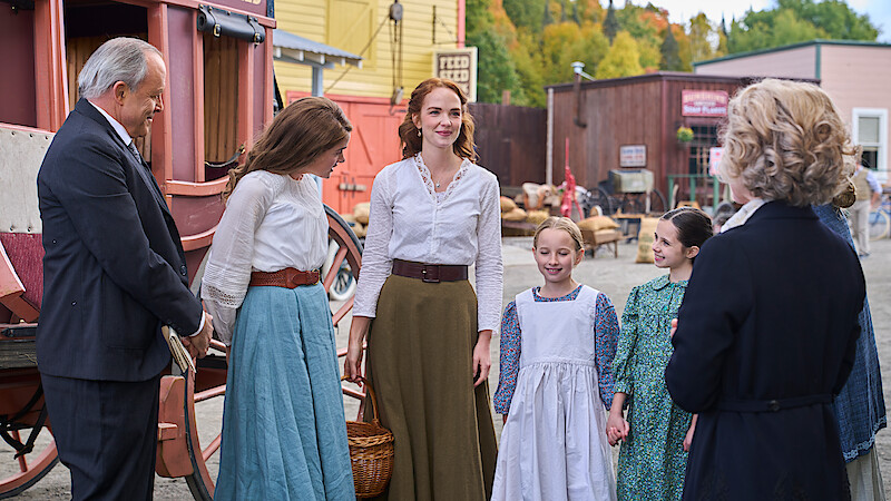 Group of people in period clothing stand and converse in a rustic, old-fashioned town setting with a stagecoach, wooden buildings, and outdoor market visible in the background.
