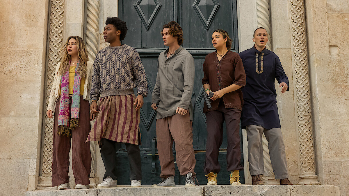 Five young adults in casual, earth-toned clothes stand together on a stone step outside an ornate building with large doors, looking alert and concerned, surrounded by intricate architectural details.