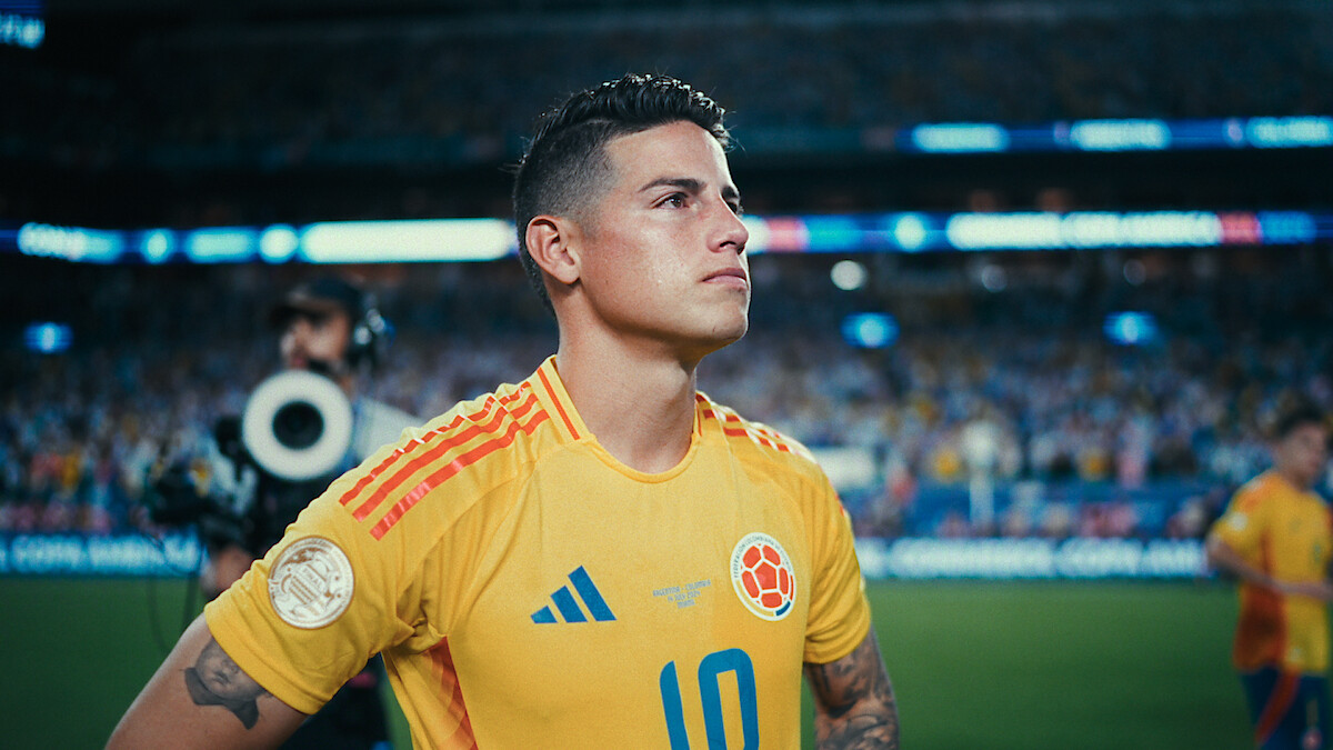 A soccer player in a yellow Colombia jersey stands focused on the field, with a stadium full of spectators and media person in the background during a match.