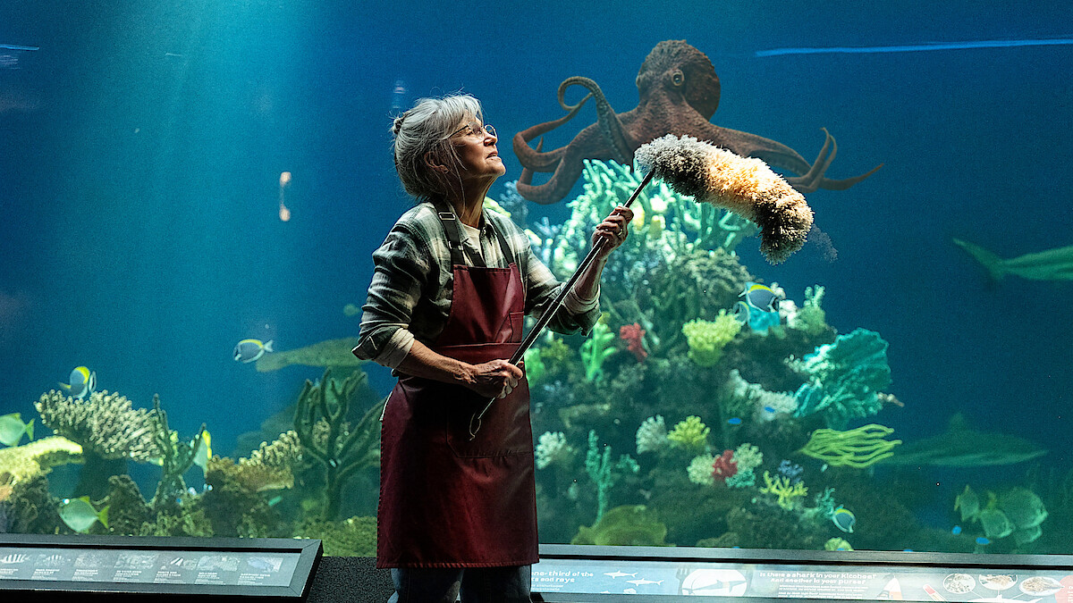 An older woman in an apron uses a duster in front of a large aquarium tank with coral, fish, and a prominent octopus visible in the water behind her, suggesting she is cleaning at an aquarium exhibit.