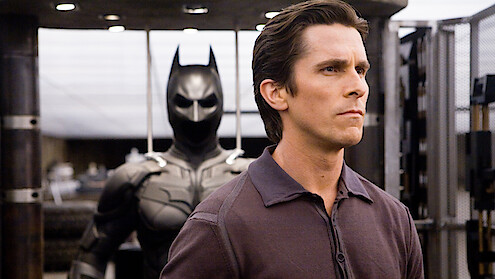 Christian Bale stands in front of Batman costume in The Dark Knight.