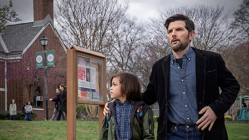 A man and child stand outside near a community notice board with a "Have You Seen Me?" poster. They look alert or concerned. The setting is a grassy area by a brick building with people and leafless trees in the background.