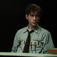 Young man in a collared shirt and tie sits in a dimly lit room, attached to a polygraph test device, with a microphone in the foreground, appearing tense or wary.