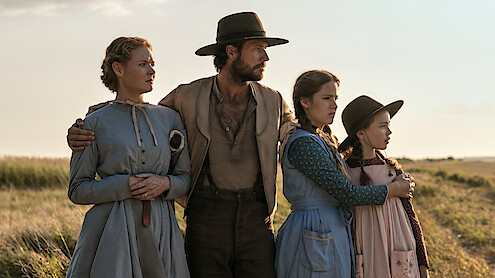 A man and three girls in old-fashioned clothing stand close together in a grassy field, looking intently into the distance under an open sky, evoking a historical or pioneer setting.