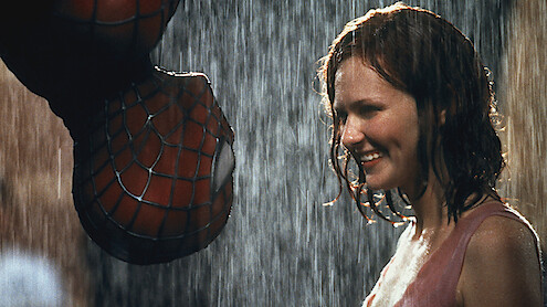 Spider-Man hangs upside down in the rain, about to kiss a smiling woman who stands close to him, both illuminated and wet, in an urban night setting.
