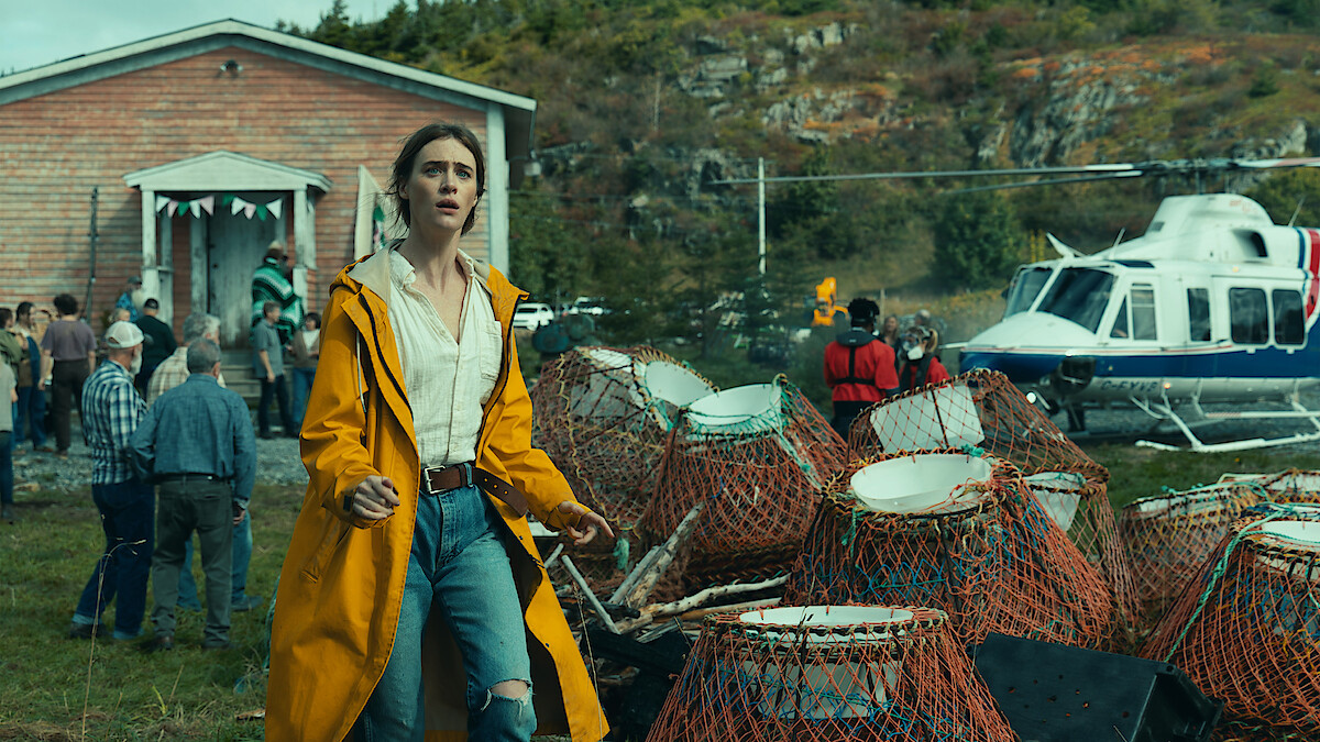 Woman in yellow raincoat stands near lobster traps, outdoors by a crowd and a helicopter, with a wooden building and forested rocky hill in the background, suggesting a coastal rural setting.