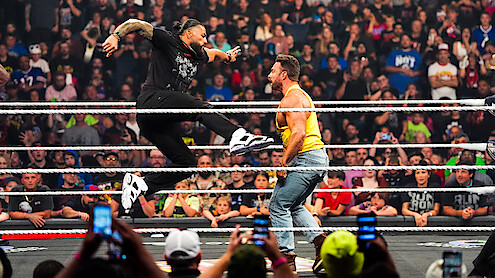 Two professional wrestlers perform a dramatic move in a wrestling ring, surrounded by cheering fans in a packed indoor arena, with several people capturing the action on their phones.
