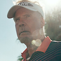 A man wearing a sports visor and striped shirt stands outdoors in the sunlight, with palm trees and blue sky in the background, giving a serious expression.