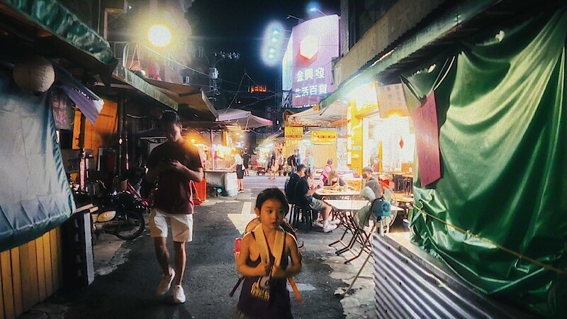 Child and adult walk through a bustling, neon-lit night market with food stalls, tables, and people dining; the scene is lively and colorful, evoking a vibrant urban atmosphere.