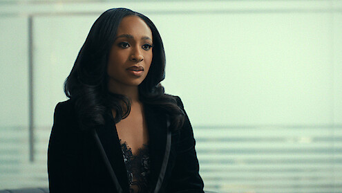 Woman with long dark hair in a black blazer sits indoors with a blurred glass wall and horizontal lines in the background, suggesting a modern office or professional setting.