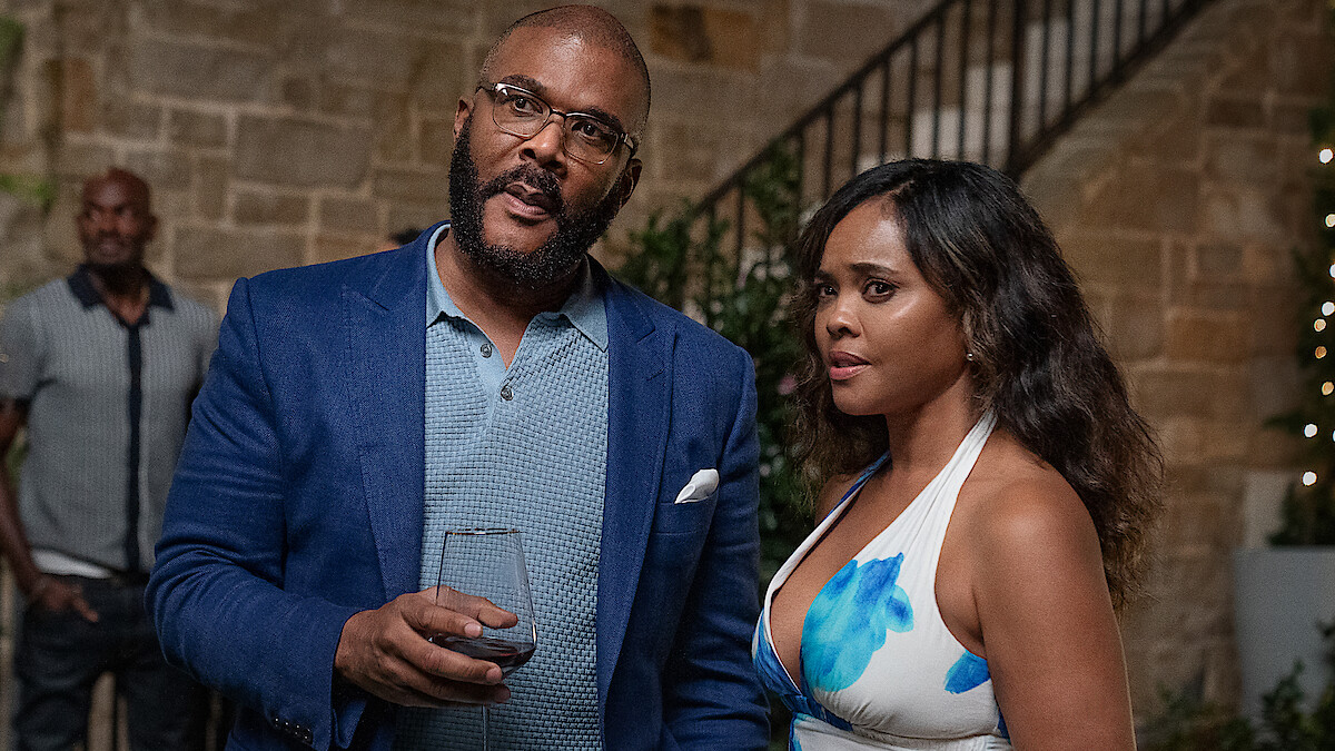 A man in a blue blazer holding a wine glass stands next to a woman in a floral dress at an upscale indoor gathering with stone walls, greenery, and a staircase in the background. Another person is blurred in the distance.