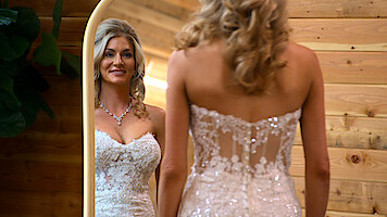 Amber Morrison in a sparkly white wedding dress looks at her reflection in a mirror. She is in a warmly lit wooden room, and her hair is styled in loose curls. The setting appears cozy and elegant.