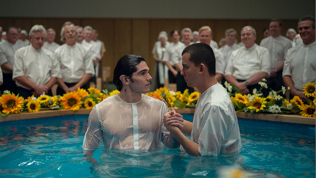 Two men in white shirts stand in a baptismal pool surrounded by sunflowers, with a group of people in white shirts and blouses watching in the background, in a brightly lit indoor setting.