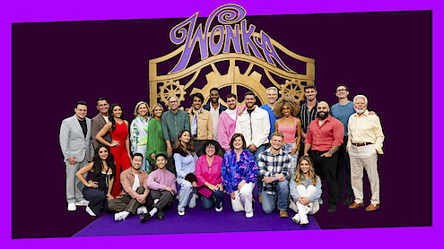 Group of people posing together in front of a large decorative "Wonka" gate backdrop, standing and sitting on a purple carpet, dressed in colorful outfits, smiling in a studio or event setting.