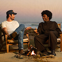 Two people sitting in wooden chairs by a campfire on a sandy beach at sunset, engaged in conversation with the ocean and pastel sky in the background.