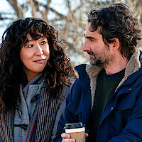 Two people sitting outdoors in warm coats, facing each other and smiling, with one holding a coffee cup. The background shows bare trees, suggesting a chilly day in a park or natural setting.