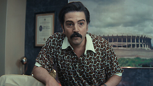 Man with dark hair and mustache in patterned shirt leans forward in an office with framed certificate on wall and large stadium photo in the background.