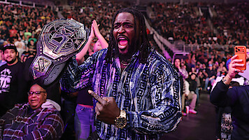 Oba Femi holding a wrestling championship belt and celebrating in a crowded arena, surrounded by fans taking photos and cheering during a live event.