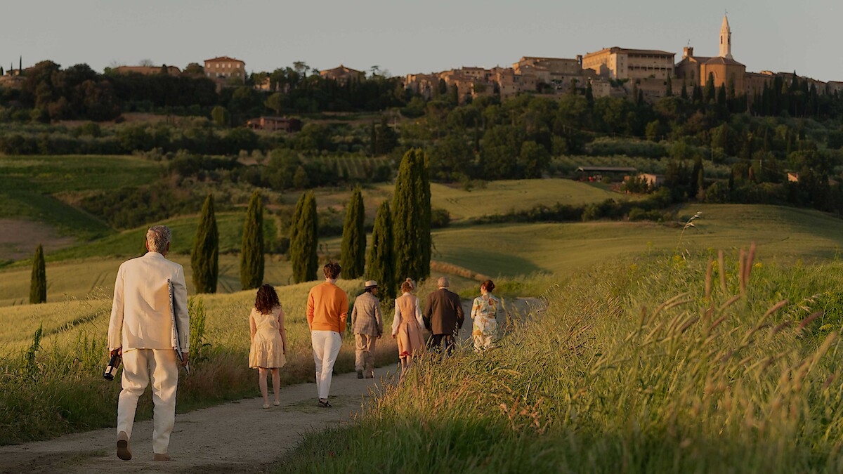 A group of people walk down a country path through green fields at sunset, heading toward a distant hilltop village, evoking a peaceful and warm atmosphere.