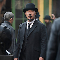 A serious man in a black pinstripe suit and bowler hat stands in an urban street scene, surrounded by others in period clothing, with green and yellow building windows in the background.