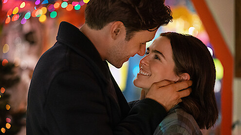 A couple embraces and smiles at each other lovingly indoors, surrounded by colorful holiday lights in a festive, warm setting.
