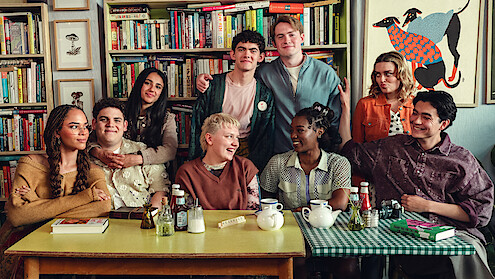 A diverse group of eight young people sitting and standing around a table in a cozy room with bookshelves and art, sharing a friendly moment, some smiling and talking, with tea and condiments on the table.