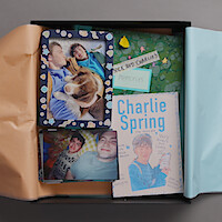A memory box contains photos of two young people, a dog, a scrapbook labeled “Nick and Charlie’s memories,” and a handmade poster for “Charlie Spring.” The box is lined with pastel tissue paper.