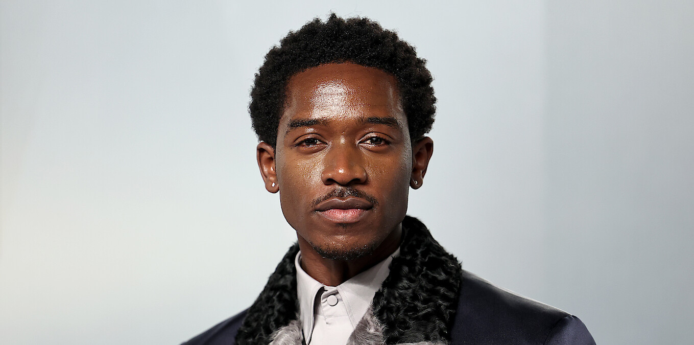 A well-dressed man with short curly hair and earrings standing in front of a plain, light-colored background, photographed in a studio setting with soft lighting.