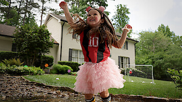 Young girl wearing a soccer jersey and pink tutu joyfully jumps in a puddle outside a suburban house, with a soccer net and trees in the background, on a cloudy day.