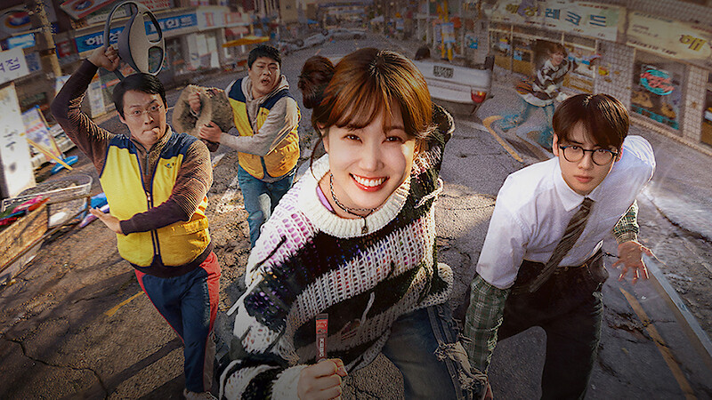 Five people, including a smiling woman in a colorful sweater and a man in glasses, run on a damaged street in a city neighborhood with cracked pavement and rundown storefronts, suggesting urgency or disaster in an urban setting.