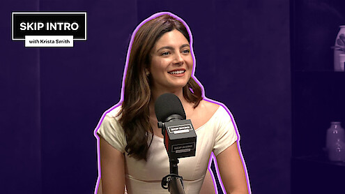 Monica Barbaro smiles in front of a microphone, framed by a deep purple background with a matching purple outline surrounding her.