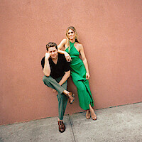 A man in a black shirt and green pants sits on a stoop next to a woman in a green dress, both posing casually against a plain pink wall in an urban outdoor setting.