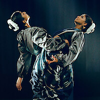 Two women in matching metallic outfits and white floral hairpieces perform a synchronized dance pose on a dark stage, creating a dramatic and artistic atmosphere with expressive movement.