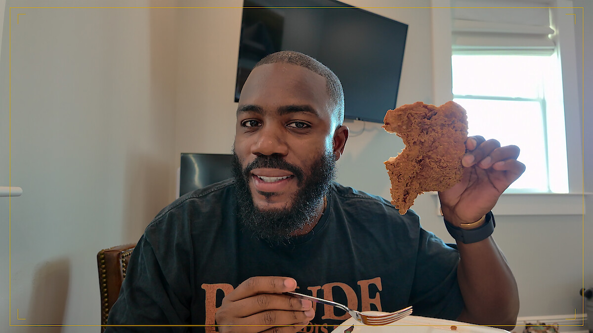 Man sitting indoors with a fork in one hand and holding a large piece of fried food in the other, smiling at the camera; modern room with a TV and window in the background.