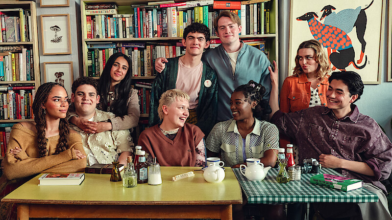 A group of eight young adults sitting and standing around tables with tea and books in a cozy room filled with bookshelves and colorful art, smiling and enjoying each other's company.