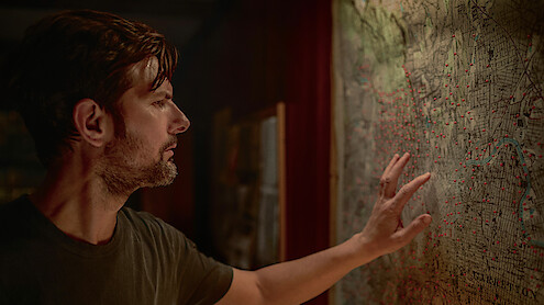 Man in dimly lit room closely examining a large, detailed map on the wall, touching the surface with his hand as he studies numerous red dots marked on the map.