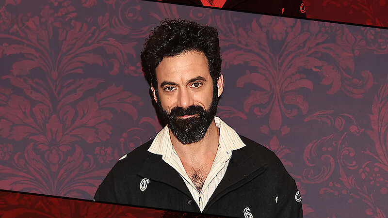 A man with dark curly hair and a full beard stands indoors against a patterned red wall, wearing a black jacket with a partially open collared shirt, posing for a portrait with a neutral expression.