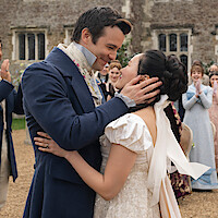 A couple dressed in Regency-era wedding attire embrace outdoors, surrounded by clapping guests in formal period clothing, with a stone building in the background.