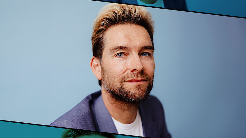 Man with blond hair and beard in a gray suit jacket and white shirt, posing against a plain light blue background in a professional portrait setting.