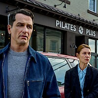 Two serious-looking people stand outside a Pilates studio on a city street, with a car parked behind them; the mood appears tense and contemplative.