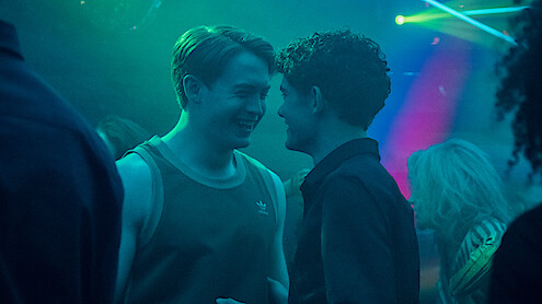 Two young men smiling closely at each other in a dimly lit nightclub with blue and green lighting, suggesting a lively social atmosphere.